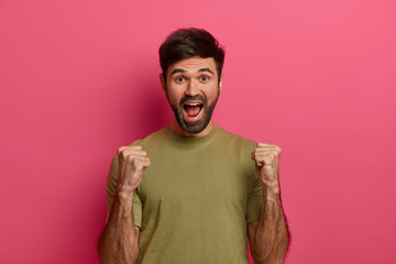 Successful teen boy raises clanched fists, celebrates triumph, looks with joy, exclaims loudly, has thick stubble, wears casual t shirt, poses over pink background screams yes, got prize, wins contest