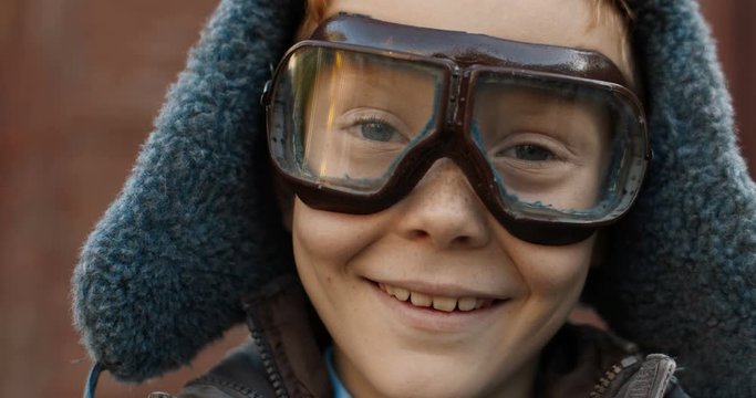 Close up of face of small Caucasian cute red-haired boy in hat and glasses smiling cheerfully to camera outdoors. Portrait of little happy child. Childhood dreams of future profession of pilot concept