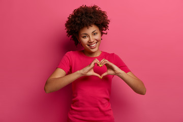 Pretty curly African American woman confesses in love, makes heart gesture, shows her true...