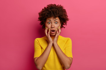 Stunned speechless young woman has shocked scared expression, keeps hands on cheeks, reacts to sudden unexpected news, wears yellow t shirt, has eyes popped out, isolated on pink background.