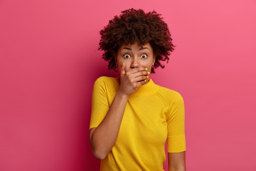 Fototapeta na wymiar Scared dark skinned woman covers mouth, holds breath from fright, stands speechless, finds out shocking truth or news, dressed in casual yellow clothes, isolated on pink background, looks worried