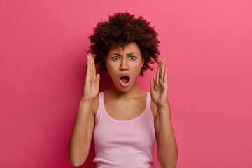 Surprised dark skinned woman makes big hand sign, demonstrates width of package, has shocked expression, dressed casually, poses over rosy background, gestures and measures something very huge