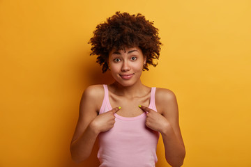 Unconfident hesitant curly haired adult woman points at herself, asks doubtfully whether she is good candidate, dressed casually, isolated over yellow background, looks friendly, asks question