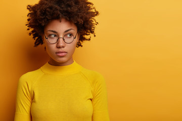 Fototapeta na wymiar Close up shot of contemplative young female model wears round spectacles ad yellow clothes, looks aside with pensive expression, thinks over plan, poses indoor, blank space for your advertisement