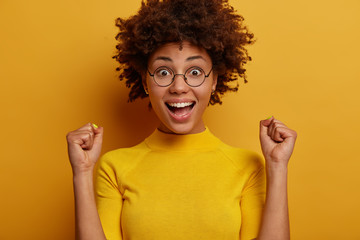 Yes, I did it! Euphoric dark skinned woman raises hands with clenched fists, looks happily, dressed in casual wear, stands against yellow background, feels like winner. People, emotions, lifestyle