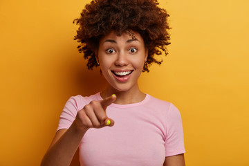 Cropped image of pretty joyful woman with toothy smile points directly at camera, sees something amazing in front, wears t shirt, has curious happy expression, isolated on yellow background.