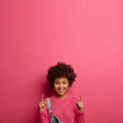 Your logo here. Pleased smiling woman with Afro hair points and promots something upwards, says follow this direction, wears knitted sweater, isolated on pink background. Marketing and advertising