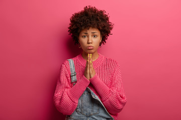 Regretful dark skinned African American woman beggs for forgiveness, feels guilty, purses lower lip, wears knitted sweater, isolated on pink background, beseeches parents for money, looks innocent
