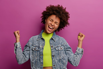 Happy relaxed woman raises clenched fists, celebrates success, tilts head, dressed in denim...