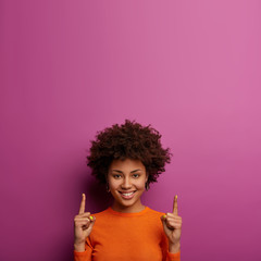 Pretty cheerful African American girl invites you going upstairs, recommends product and points above, has happy expression, wears casual orange jumper, isolated on purple wall, gazes with joy