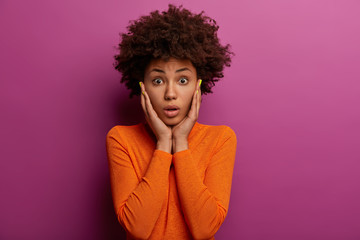 Fototapeta na wymiar Fascinated Afro American woman grabs cheeks, dressed in orange jumper, realizes she forgot about something important, receives big fine, poses against purple background. Human face expressions