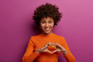 I love you with all my heart. Smiling dark skinned woman makes heart gesture, falls in love with someone, tells I am fully yours, has friendly look, dressed casually, isolated on purple background