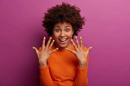 Curly ethnic woman shows manicured yellow nails, has glad expression, smiles happily, glad after visiting manicurist, wears casual orange jumper, isolated over purple background, keeps hands raised