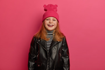 Positive smiling red head girl giggles positively, wears pink hat and leather jacket, looks gladfully at camera, isolated over pink background. Children, style concept. Lovely daughter enjoys shopping