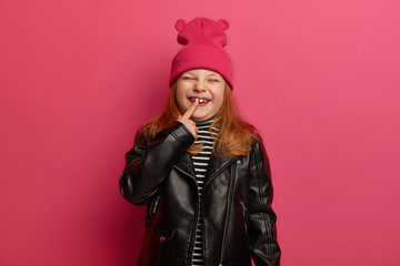 Happy little ginger girl in fashionabe clothes, indicates at her new tooth, has unforgettable childhood, squints face with pleasure, poses against briht pink background. Chidren growth concept