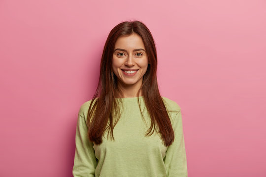 Portrait of attractive woman has healthy skin, has toothy smile, looks directly at camera, wears green jumper, has long straight hair, poses against pink pastel background. Face expressions concept