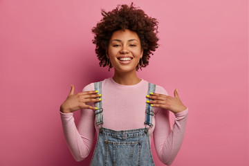 Positive young African American woman points at herself, feels proud, smiles broadly, being in high spirit, dressed in casual wear, poses against pink pastel background, has confident expression