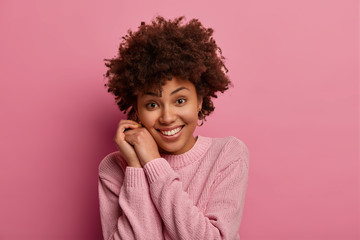 Fototapeta na wymiar Photo of pretty young tender woman with Afro hair smiles gently, keeps hands near face, looks directly at camera, wears casual sweater, hears somethning good, poses against pink studio background