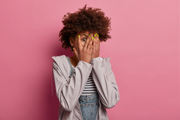 Fototapeta na wymiar Omg, how scarying! Frightened curly haired woman peeks through fingers, hides face with palms, looks with scared expression, doesnt want to see something awful, wears grey jacket isolated on pink wall
