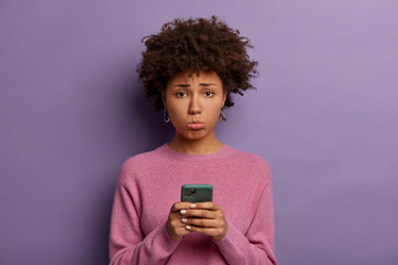 Unhappy disappointed woman with Afro hair, purses lower lip, holds smartphone, sad to miss chance of good shopping sale, upset not to receive call from boyfriend, poses indoor, dressed casually - obrazy, fototapety, plakaty