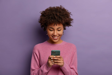 Pretty curly woman holds modern mobile phone, types messages on smartphone device, enjoys online...