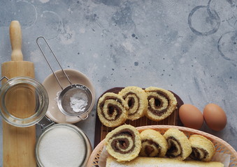 Pastry background.Homemade roll with plum jam, sprinkled with powdered sugar, on a gray wooden background with ingredients for cooking, space for text.