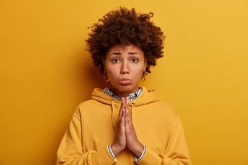 Fototapeta na wymiar Let me please. Sad pleading African American woman asks for permission, holds hands in pray, says forgive me, poses against yellow background, wears sweatshirt. Begging and saying forgive me.
