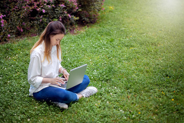 young girl surfing the internet, a student sitting in a park on green grass and typing on her laptop, online study and work, freelance