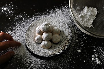 Shortbread balls on a dark plate with a striped pattern around the rim and dusted with icing sugar together with a sieve holding icing sugar all against a dark wooden background