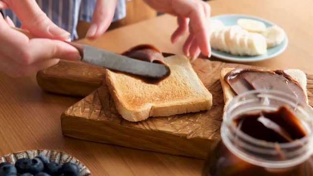 Woman spreading chocolate nut butter on toasted bread. Process of cooking tasty breakfast or lunch sandwich with chocolate nut butter