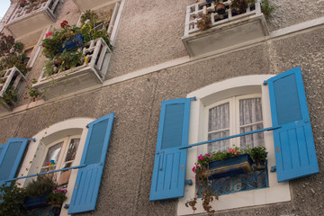 Fototapeta na wymiar Beautiful medieval building with flowers and blue window shutters. Vintage facade of mediterranean houses. Street in old town in Europe. Stone wall with small balconies and flowers. 