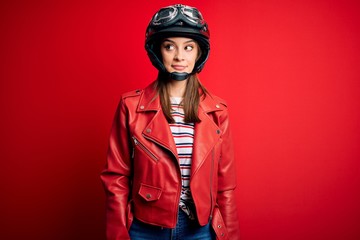 Young beautiful brunette motocyclist woman wearing motorcycle helmet and red jacket smiling looking to the side and staring away thinking.