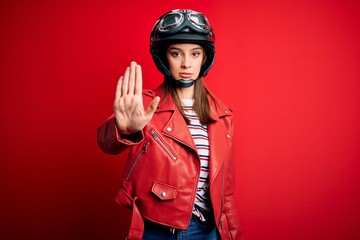 Young beautiful brunette motocyclist woman wearing motorcycle helmet and red jacket doing stop sing with palm of the hand. Warning expression with negative and serious gesture on the face.