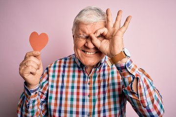 Naklejka premium Senior handsome hoary romantic man holding red paper heart shape over pink background with happy face smiling doing ok sign with hand on eye looking through fingers