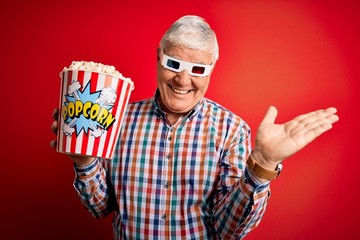Senior hoary man watching film using 3d glasses eating popcorn over red background very happy and excited, winner expression celebrating victory screaming with big smile and raised hands