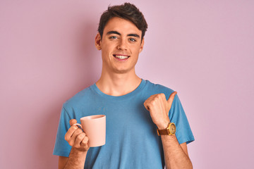 Teenager boy drinking a cup of coffee over isolated pink background pointing and showing with thumb...