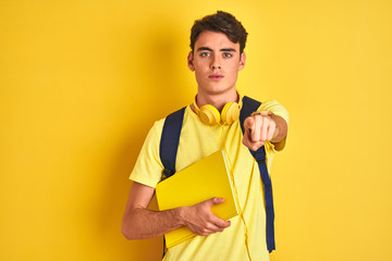Teenager boy wearing headphones and backpack reading a book over isolated background pointing with...