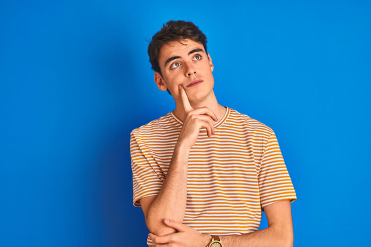 Teenager boy wearing casual t-shirt standing over blue isolated background with hand on chin thinking about question, pensive expression. Smiling with thoughtful face. Doubt concept.