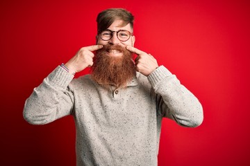 Handsome Irish redhead man with beard wearing casual sweater and glasses over red background Smiling with open mouth, fingers pointing and forcing cheerful smile