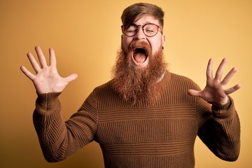 Handsome Irish redhead man with beard wearing glasses and winter sweater over yellow background...
