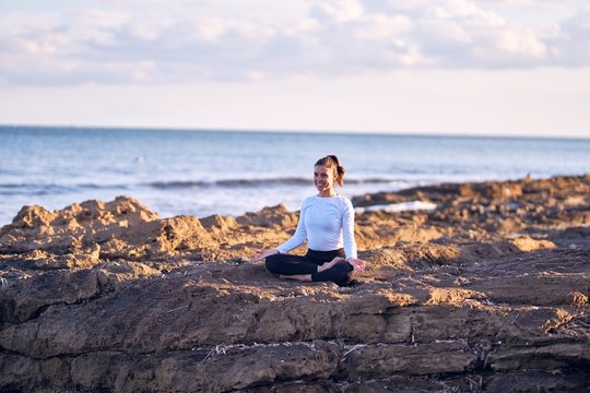 Young beautiful sportwoman smiling happy practicing yoga. Coach with smile on face teaching lotus pose at the beach