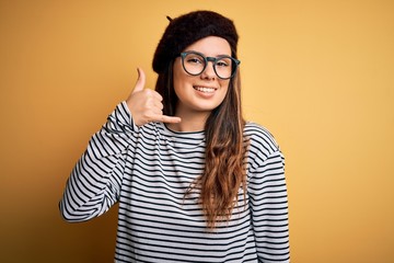 Young beautiful brunette woman wearing french beret and glasses over yellow background smiling doing phone gesture with hand and fingers like talking on the telephone. Communicating concepts.