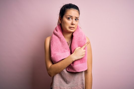 Young beautiful brunette sportswoman wearing sportswear and towel over pink background Pointing aside worried and nervous with forefinger, concerned and surprised expression