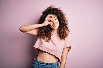 Young beautiful woman with curly hair and piercing wearing casual pink t-shirt doing ok gesture with hand smiling, eye looking through fingers with happy face.