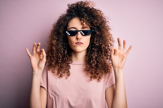 Young beautiful woman with curly hair and piercing wearing funny thug life sunglasses relax and smiling with eyes closed doing meditation gesture with fingers. Yoga concept.
