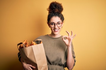 Young beautiful woman with curly hair and piercing holding paper bag with croissants doing ok sign...