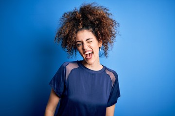 Fototapeta na wymiar Young beautiful woman with curly hair and piercing wearing casual blue t-shirt winking looking at the camera with sexy expression, cheerful and happy face.