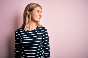 Fototapeta premium Young beautiful blonde woman wearing casual striped sweater over pink isolated background looking away to side with smile on face, natural expression. Laughing confident.