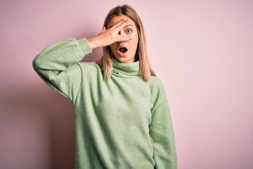 Young beautiful blonde woman wearing winter wool sweater over pink isolated background peeking in shock covering face and eyes with hand, looking through fingers with embarrassed expression.