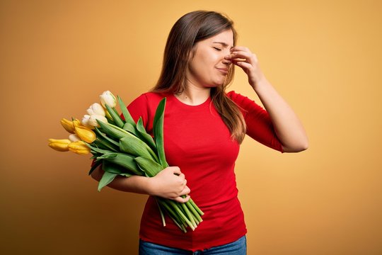 Young Blonde Woman Holding Romantic Bouquet Of Tulips Flowers Over Yellow Background Smelling Something Stinky And Disgusting, Intolerable Smell, Holding Breath With Fingers On Nose. Bad Smell